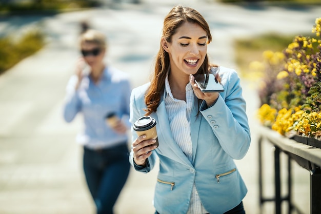 Jovem empresária sorridente está andando na frente do escritório e enviando mensagem de voz durante a pausa para o café.