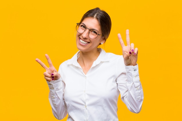 Jovem empresária sorrindo e olhando feliz, amigável e satisfeito, gesticulando vitória ou paz com as duas mãos contra a parede laranja