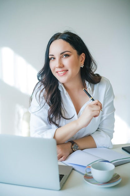 Jovem empresária trabalha no computador Uma linda mulher de cabelos escuros em uma camisa branca trabalha online em um escritório brilhante Bebendo café e comendo um croissant