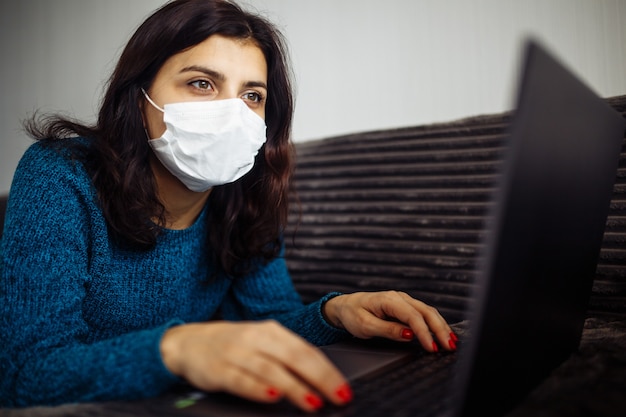 Jovem empresária trabalhando em casa durante a quarentena devido à pandemia de coronavírus. Linda garota fica em casa usando máscara médica e digitando em um laptop. Conceito mundial de epidemia Covid-19.