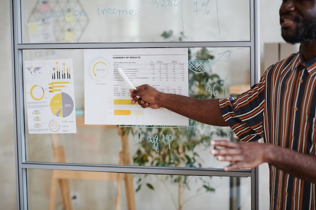 Foto jovem empresário africano apontando para um documento financeiro na parede de vidro e apresentando-o aos colegas no escritório