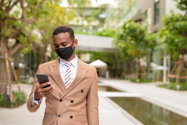 Jovem empresário africano com máscara usando telefone na cidade com a natureza