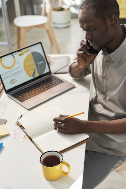 Jovem empresário africano falando no celular e fazendo anotações em seu caderno no escritório