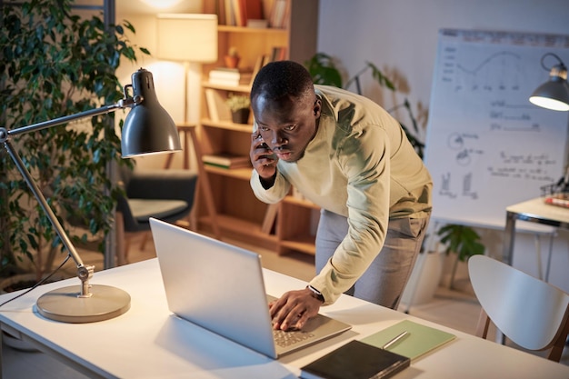 Jovem empresário africano usando laptop na mesa enquanto fala no celular com o colega que trabalha até tarde no escritório