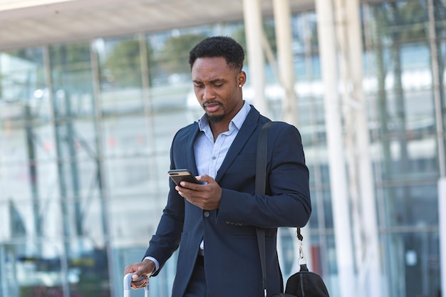 Jovem empresário afro-americano confiante e bem-sucedido, movendo-se com o celular nas mãos contra um prédio de escritórios moderno, um homem de negócios preto no centro da cidade anda usando um telefone celular em um terno formal