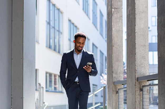 Jovem empresário afro-americano confiante e bem-sucedido, movendo-se com o celular nas mãos contra um prédio de escritórios moderno, um homem de negócios preto no centro da cidade anda usando um telefone celular em um terno formal