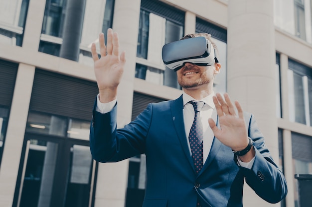 Jovem empresário alegre com barba, vestido com um terno azul formal, experimentando óculos de realidade virtual, verificando a realidade virtual, explorando o mundo digital, sozinho ao lado de um prédio no fundo