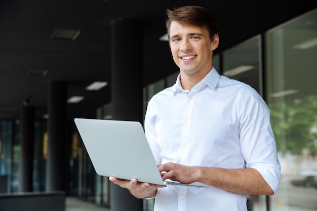 Jovem empresário alegre segurando um laptop e falando no celular, perto do centro de negócios