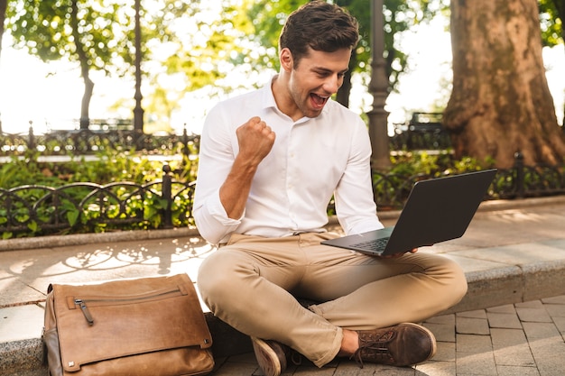Jovem empresário alegre sentado ao ar livre, trabalhando em um laptop, comemorando o sucesso