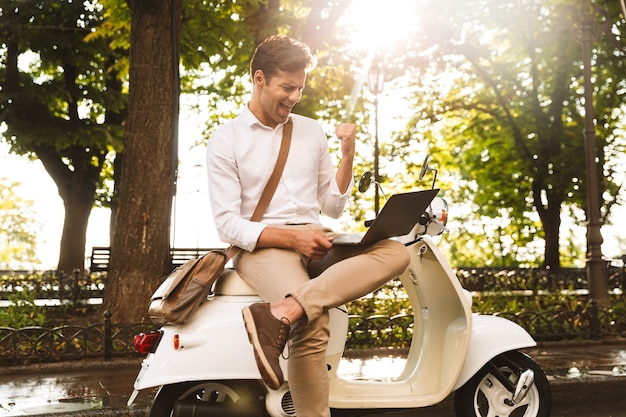 Jovem empresário alegre sentado em uma moto ao ar livre, trabalhando em um laptop