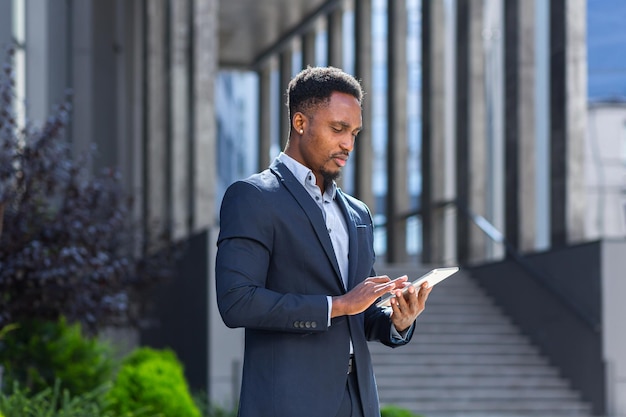 Jovem empresário americano africano em terno de negócio formal em pé trabalhando com tablet em mãos no prédio de escritórios moderno do fundo do lado de fora. homem usando smartphone ou telefone celular ao ar livre na rua da cidade