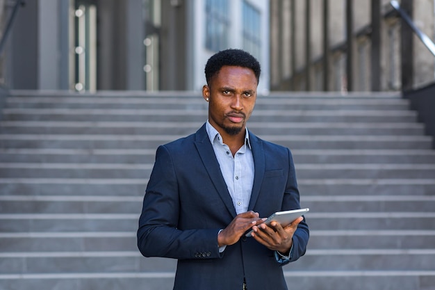 Jovem empresário americano africano em terno de negócio formal em pé trabalhando com tablet em mãos no prédio de escritórios moderno do fundo do lado de fora. homem usando smartphone ou telefone celular ao ar livre na rua da cidade