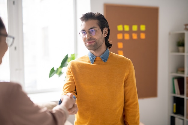 Jovem empresário amigável de óculos e roupas casuais cumprimentando seu novo parceiro ou colega com um aperto de mão no escritório