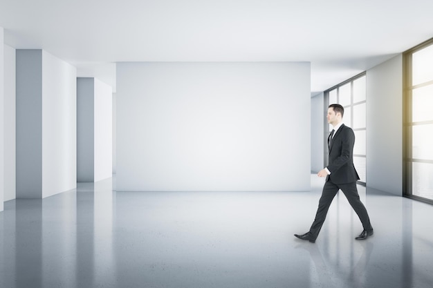 Jovem empresário andando em um quarto minimalista e espaçoso com paredes claras em branco e janelas para o chão Mockup