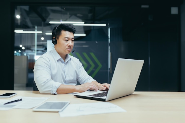 Jovem empresário asiático bonito trabalhando no laptop em fones de ouvido com microfone sentado em uma mesa