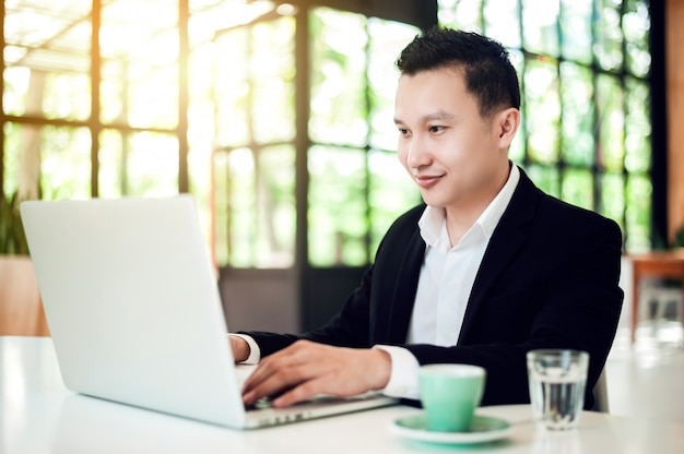 Jovem empresário asiático de terno sentado à mesa no café usando o laptop com xícara de café e olhando para a tela do laptop.