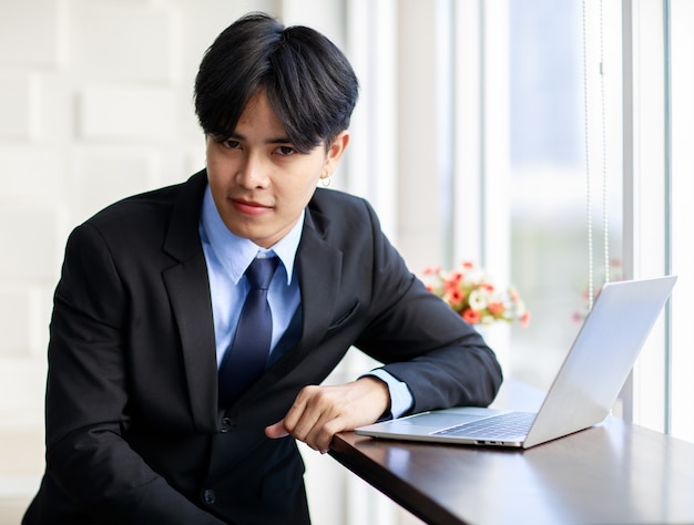 Jovem empresário asiático é inteligente e bonito usando terno preto, sentado enquanto digita no teclado no laptop perto da janela no escritório de manhã.