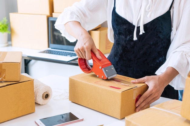 Jovem empresário asiático selando uma caixa com fita adesiva na mesa. preparando-se para o transporte, embalagem, on-line, venda, comércio eletrônico, wotk no / do conceito de casa. fechar-se.