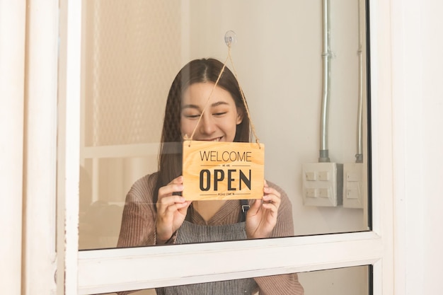 Foto jovem empresário asiático sorridente, funcionário de varejo, mulher de cafeteria, garota virando, definindo a placa de sinalização para abrir para o cliente de boas-vindas, reabrir a loja após fechar a quarentena de bloqueio no conceito covid.label.