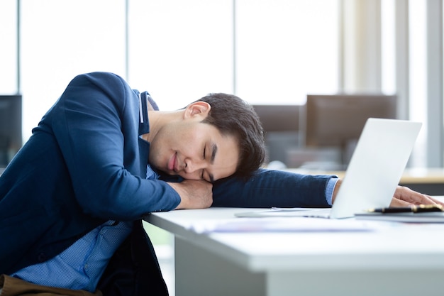 Jovem empresário asiático trabalhou até tarde e adormeceu no computador portátil no fundo da sala de escritório.