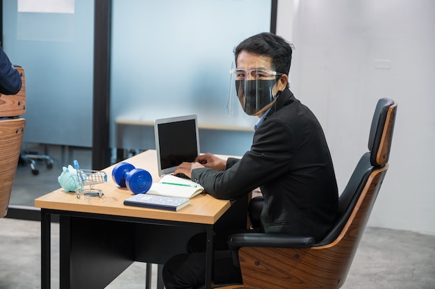 Jovem empresário asiático usando protetor facial, olhando para a câmera e trabalhando com laptop e cofrinho, haltere, carrinho e notebook na mesa do escritório