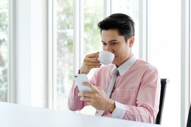 Jovem empresário asiático usando telefone inteligente e tomando café no escritório