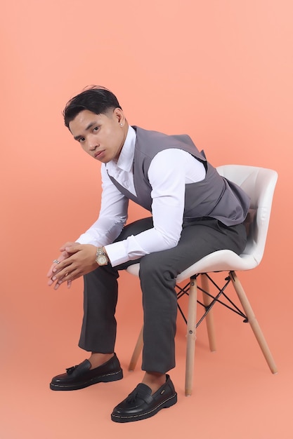 Jovem empresário asiático vestindo um colete de terno olha para a câmera sentada em uma cadeira branca