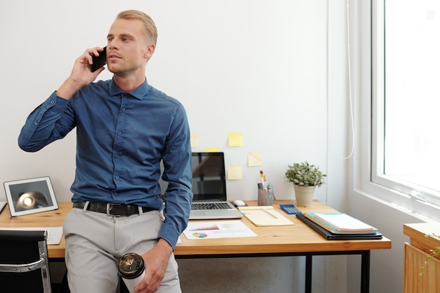 Jovem empresário atraente com uma xícara de café descartável encostado na mesa do escritório e falando ao telefone com um parceiro de negócios ou cliente