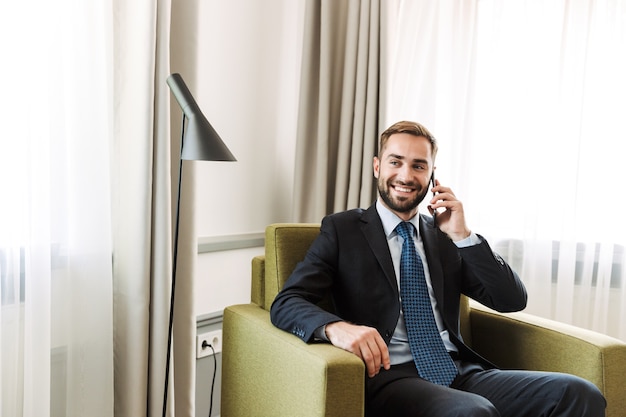 Jovem empresário atraente de terno sentado em uma cadeira no quarto do hotel