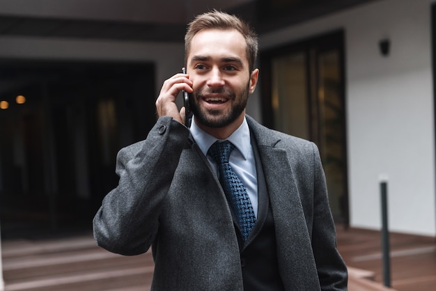 Jovem empresário atraente vestindo terno caminhando ao ar livre, usando telefone celular, conversando