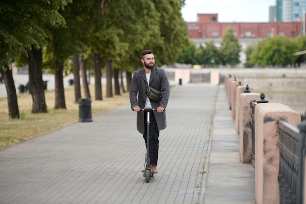 Jovem empresário barbudo contemporâneo andando de casaco em uma scooter elétrica em ambiente urbano à beira do rio pela manhã