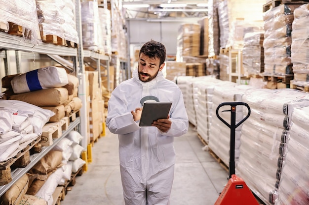 Jovem empresário barbudo dedicado em uniforme estéril branco protetor, andando pelo armazém e usando o tablet para verificar o salário das mercadorias. conceito de surto do vírus corona / covid 19.