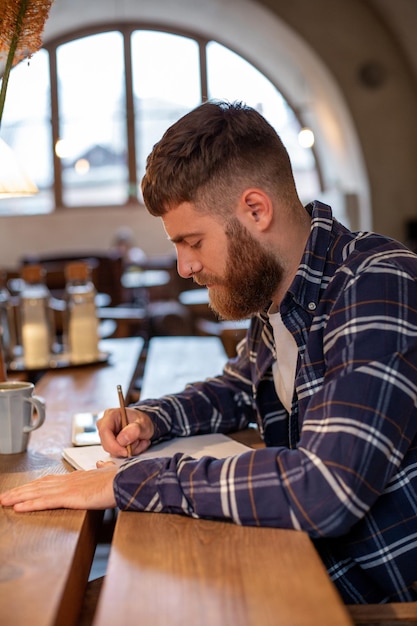 Jovem empresário barbudo se senta em um café em casa à mesa e escreve no caderno perto de computadores tablet de mentiras.