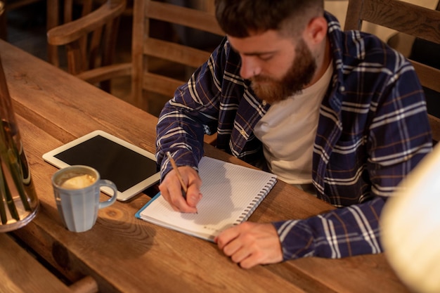 Jovem empresário barbudo se senta em um café em casa à mesa e escreve no caderno perto de computadores tablet de mentiras.