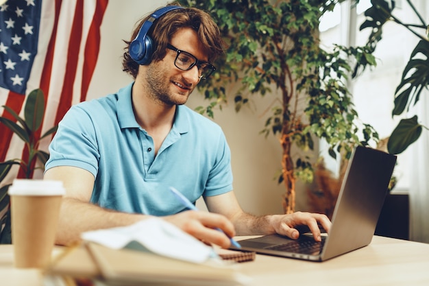 Jovem empresário barbudo sentado à mesa com o laptop e a bandeira americana