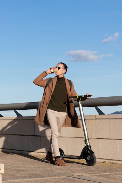 Jovem empresário bebendo café em uma xícara enquanto percorre a cidade com sua scooter elétrica sob um céu azul