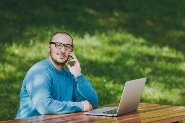 Jovem empresário bem sucedido sorridente homem inteligente ou estudante em casual camisa azul, óculos, sentado à mesa, falando no celular no parque da cidade usando laptop, trabalhando ao ar livre. Conceito de escritório móvel.