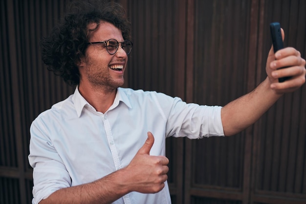 Jovem empresário bonitão tirando autoretrato com seu telefone inteligente ao ar livre Homem sorridente feliz vestindo camisa branca e óculos redondos tomando selfie contra a parede na rua