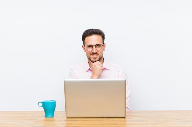 Jovem empresário bonito olhando feliz e sorrindo com a mão no queixo, pensando ou fazendo uma pergunta, comparando opções