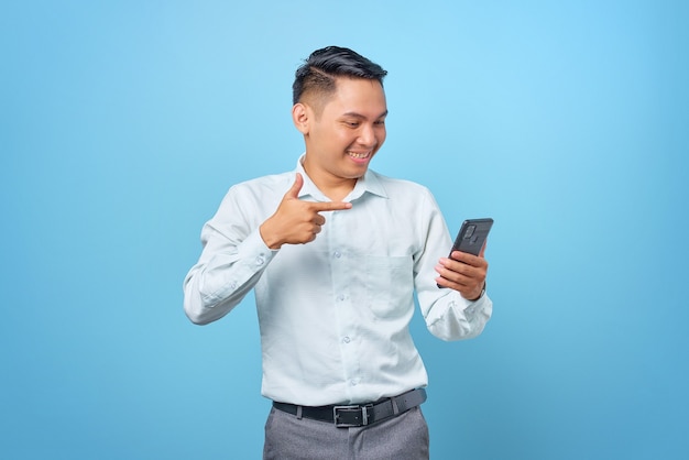 Jovem empresário bonito sorridente apontando o dedo para o smartphone sobre fundo azul