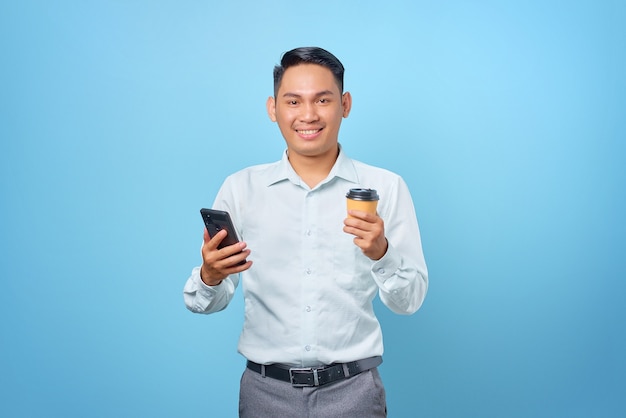 Jovem empresário bonito sorridente segurando um smartphone e uma xícara de café sobre fundo azul