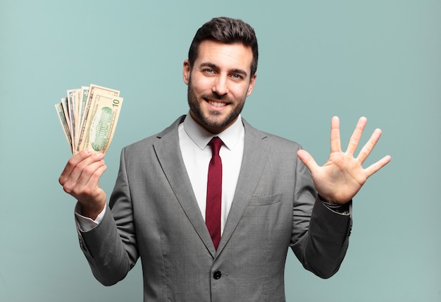 Jovem empresário bonito sorrindo e parecendo amigável, mostrando o número cinco ou quinto com a mão para a frente