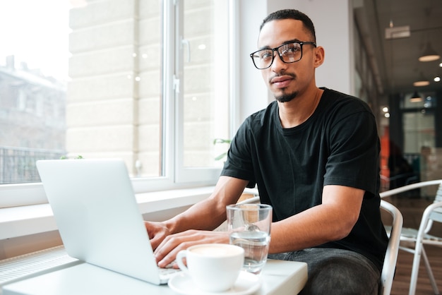 jovem empresário bonito usando óculos, sentado perto da janela e digitando no laptop
