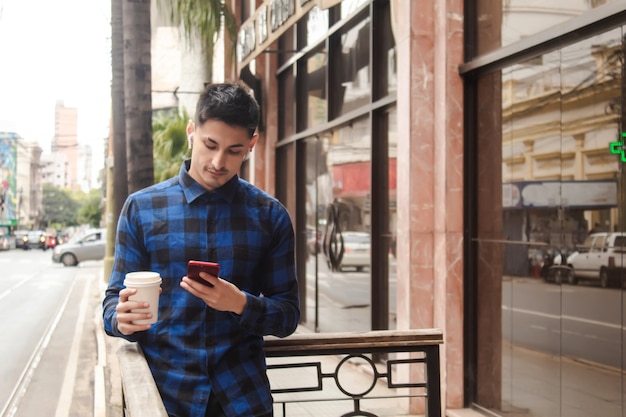 Jovem empresário bonito usando seu telefone inteligente, enquanto segura um café com a outra mão.