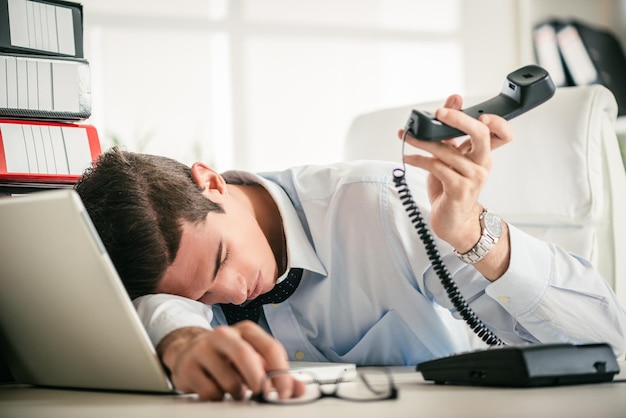 Foto jovem empresário cansado dormindo na mesa do escritório com um telefone na mão na frente do laptop e um monte de fichários.