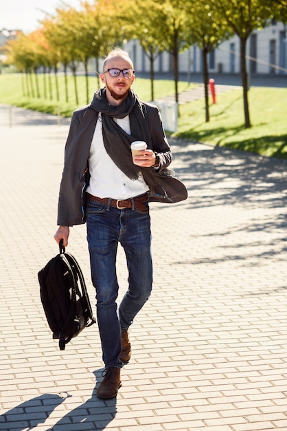 Jovem empresário caucasiano com mochila e xícara de café ao ar livre perto de centro de negócios moderno.