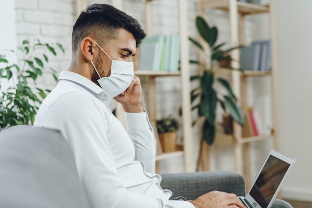 Jovem empresário com máscara facial sentado no sofá e usando o laptop em casa