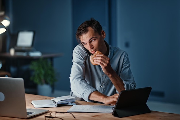 Jovem empresário comendo um hambúrguer enquanto trabalhava em um novo projeto