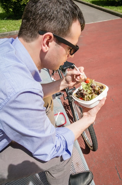 Jovem empresário comendo uma salada na hora do almoço, sentado em um banco ao ar livre