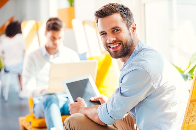 Jovem empresário confiante. Jovem feliz segurando um tablet digital e olhando para a câmera enquanto seus colegas trabalham em segundo plano